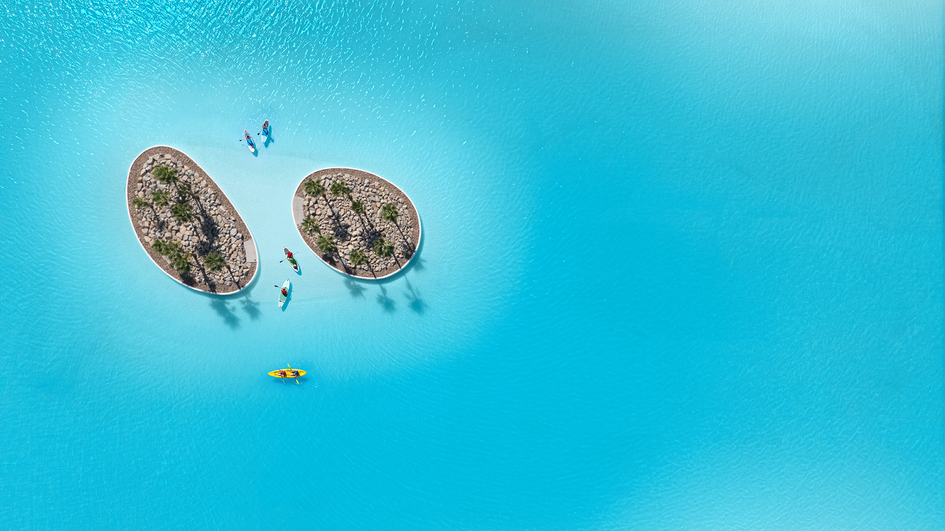Aerial view of kayakers and paddleboarders around two palm tree islands in the turquoise water along the Cotino lagoon shoreline, Crystal Lagoons lagoon in Rancho Mirage, California